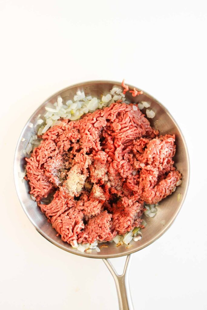 A stainless steel pan with raw ground beef, chopped onions, and various spices&mdash;perfect for making flavorful Cowboy Sliders&mdash;on a white background.