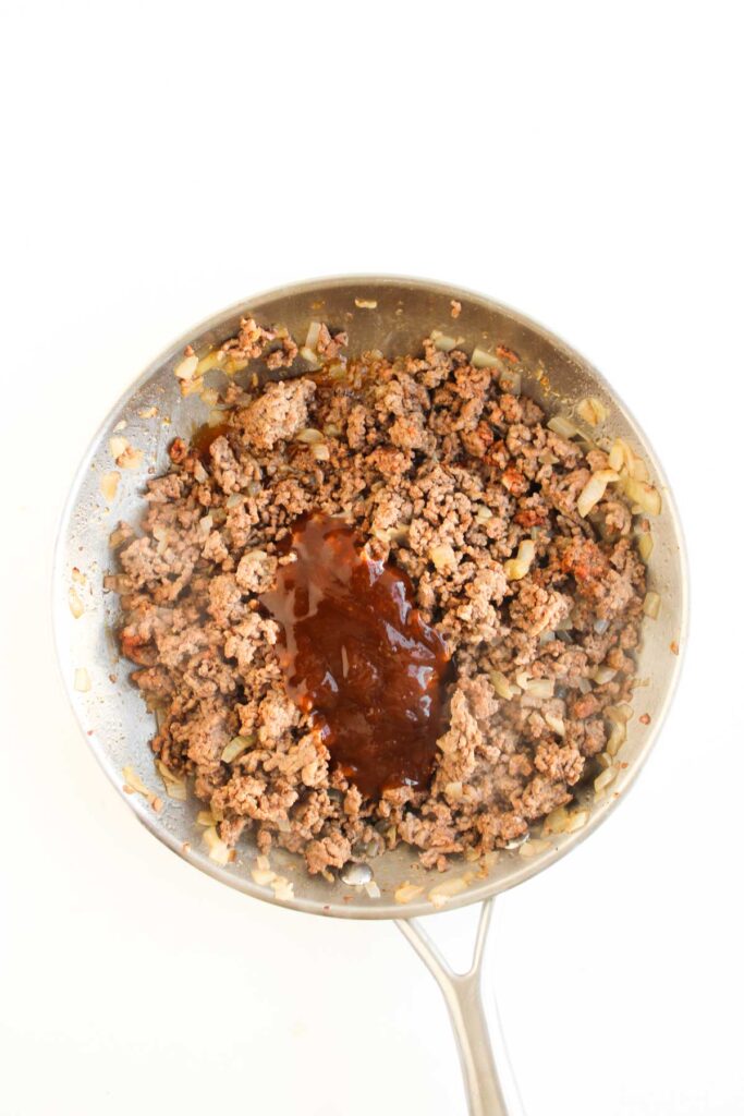 A skillet filled with cooked ground beef, chopped onions, and a dollop of dark sauce in the center, perfect for making Cowboy Sliders, photographed from above on a white background.
