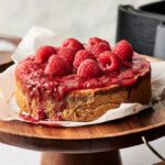 An Air Fryer Raspberry Cheesecake topped with raspberry sauce and fresh raspberries sits on parchment paper atop a wooden cake stand.