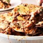 A close-up of a serving of beef moussaka with layers of eggplant, ground meat, and creamy sauce being lifted from a white casserole dish.