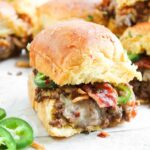 A close-up of a Cowboy Slider featuring ground beef, melted cheese, bacon, and sliced jalapeños on a soft bun, with more Cowboy Sliders and jalapeño slices in the background.