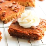 A slice of Pumpkin Pecan Pie Bar topped with a swirl of cream cheese frosting sits on a cooling rack, with more Pumpkin Pecan Pie Bars in the background.