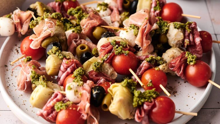 A plate of skewers with cherry tomatoes, olives, mozzarella balls, artichoke hearts, cured meats, and green pesto.