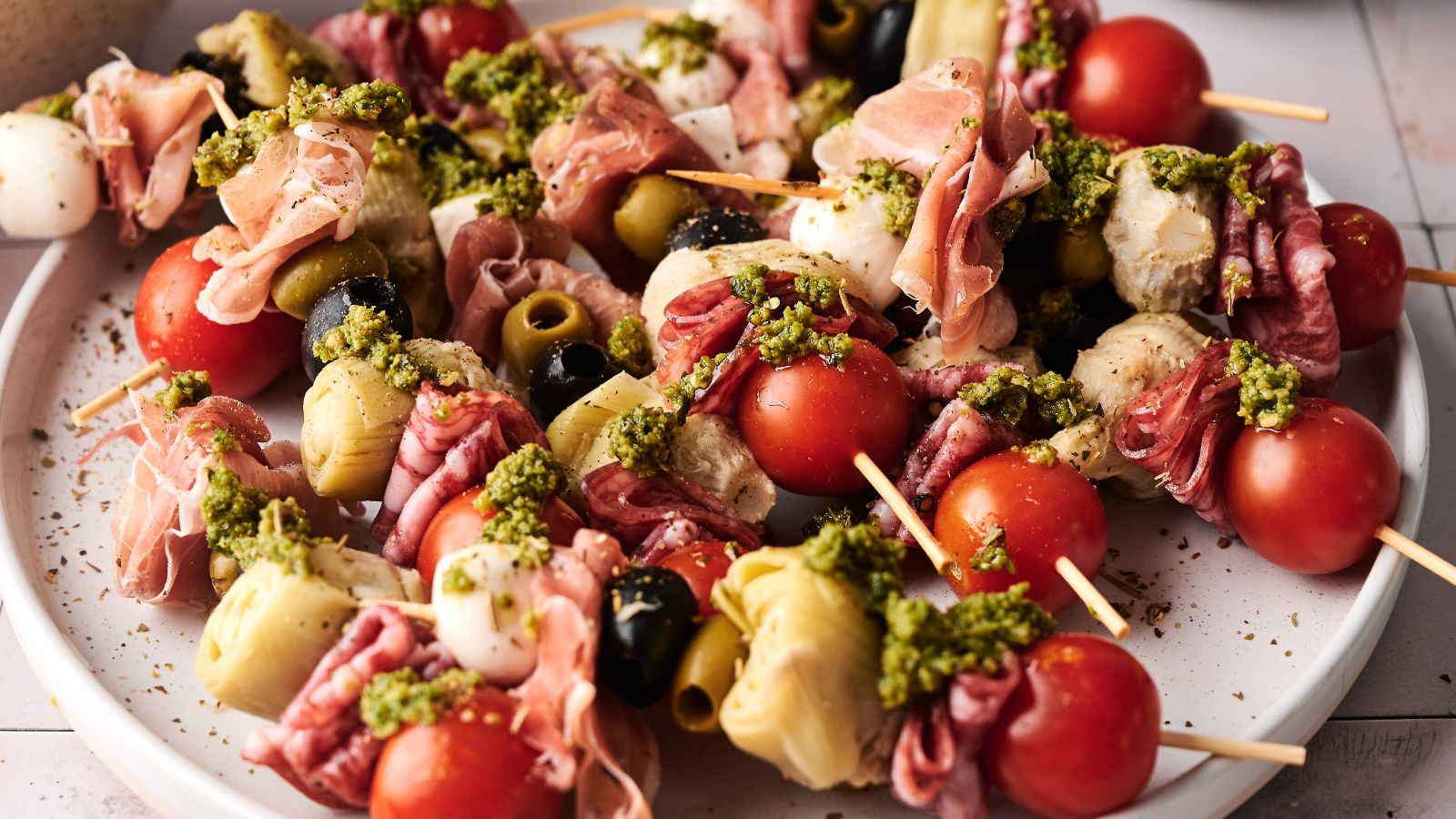 A plate of antipasto skewers with cherry tomatoes, mozzarella, olives, artichoke hearts, cured meats, and a drizzle of green pesto.