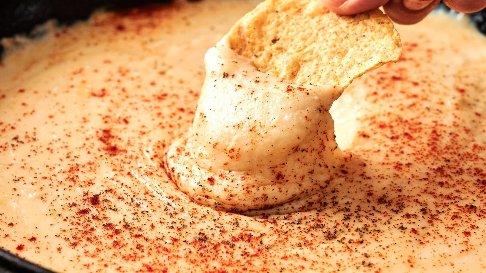 A hand dips a tortilla chip into creamy cheese dip sprinkled with paprika and black pepper.