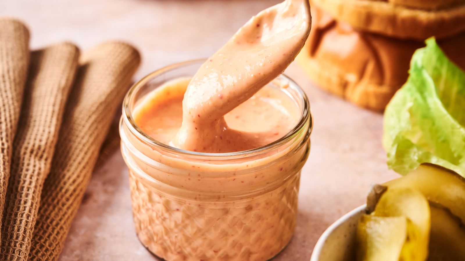 A small glass jar filled with creamy pink sauce, with a spoon lifting out some sauce. Lettuce, pickles, and napkins are visible nearby.