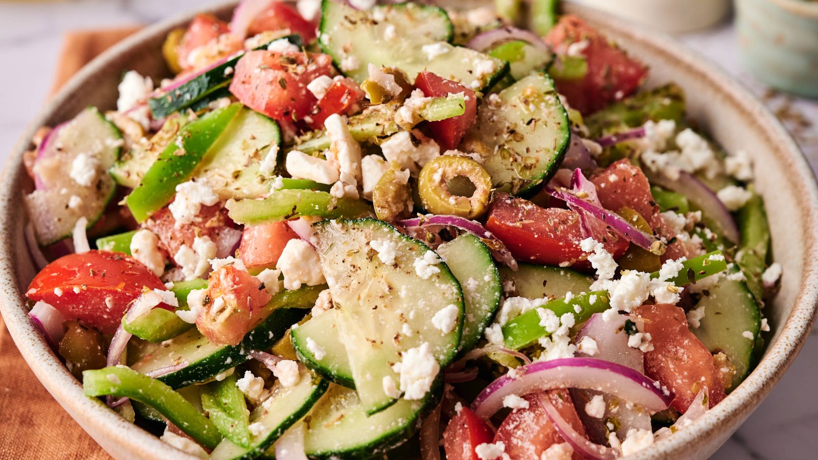 A bowl of Greek salad with sliced cucumbers, tomatoes, red onions, green bell peppers, olives, and crumbled feta cheese.