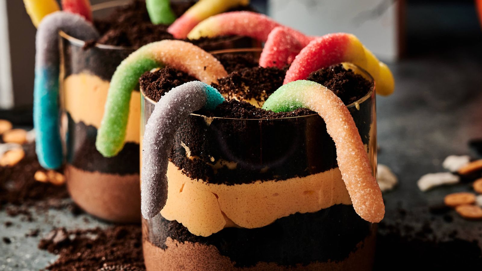 A close-up of dessert cups layered with chocolate, cream, crushed cookies, and topped with colorful gummy worms.