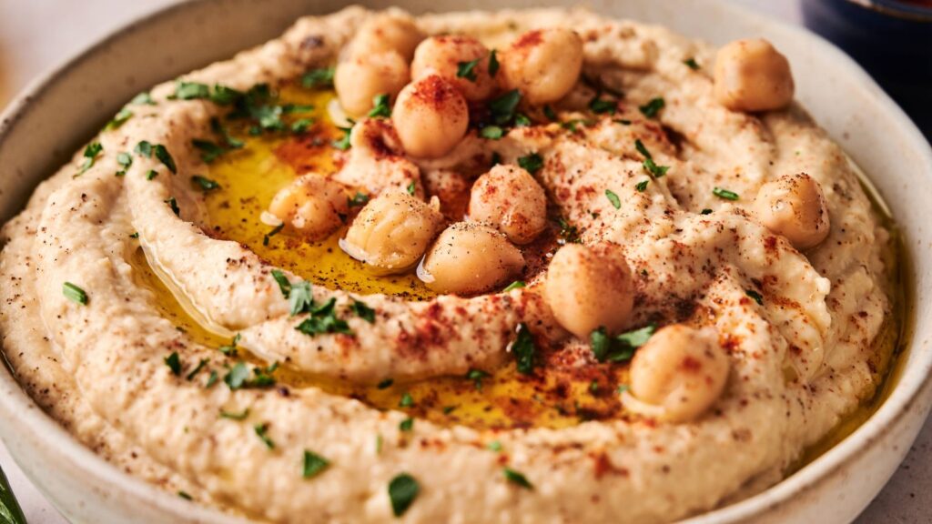 A bowl of hummus topped with whole chickpeas, olive oil, chopped parsley, and paprika.