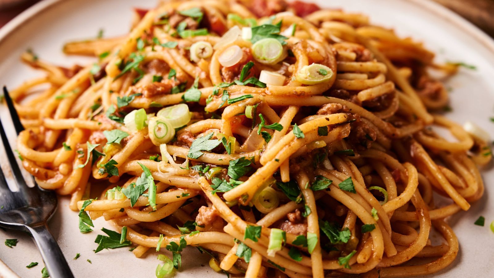 A plate of spaghetti with tomato sauce, ground meat, chopped green onions, and parsley, served with a black fork.