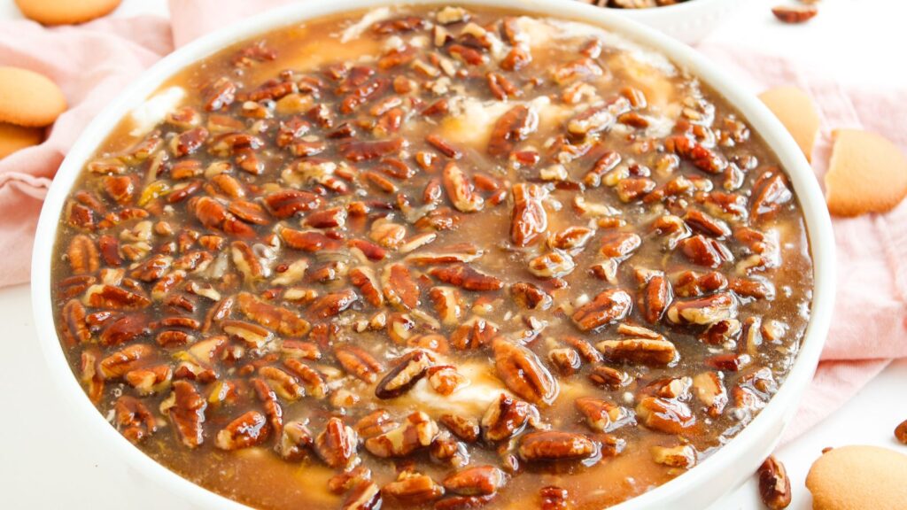 A close-up of a round dish filled with banana pudding topped with a glossy layer of chopped pecans in caramel sauce, surrounded by vanilla wafer cookies.