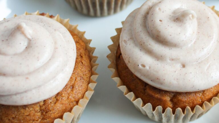 Two cupcakes with light brown frosting in paper liners on a white surface.