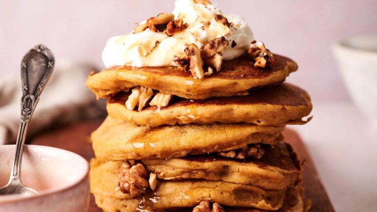 A stack of pancakes topped with whipped cream, chopped walnuts, and drizzle of syrup, with a spoon and bowl beside them.