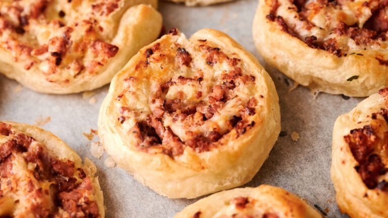 Close-up of several baked puff pastry pinwheels filled with minced meat and cheese on a parchment-lined baking tray.