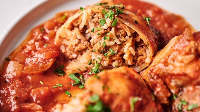 Close-up of stuffed cabbage rolls filled with rice and meat, topped with tomato sauce and chopped parsley on a white plate.
