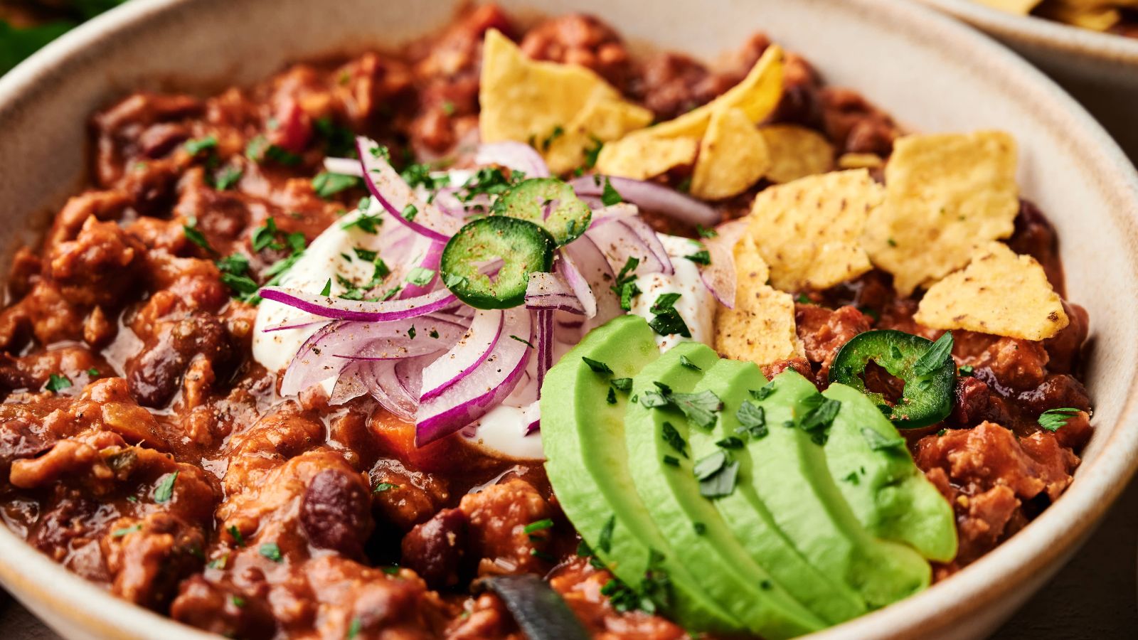 A bowl of chili topped with sliced avocado, red onions, jalapeños, sour cream, chopped herbs, and tortilla chips.
