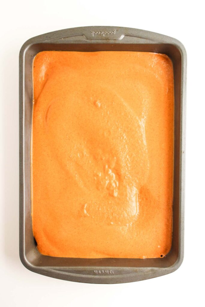 A rectangular metal baking pan filled with an even layer of orange Pumpkin Pecan Pie Bars batter, viewed from above on a white background.