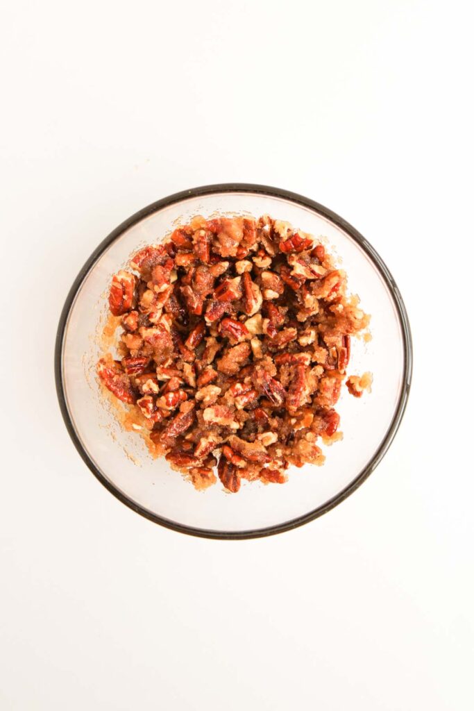 A glass bowl filled with chopped candied pecans, perfect for topping Pumpkin Pecan Pie Bars, sits on a white background.