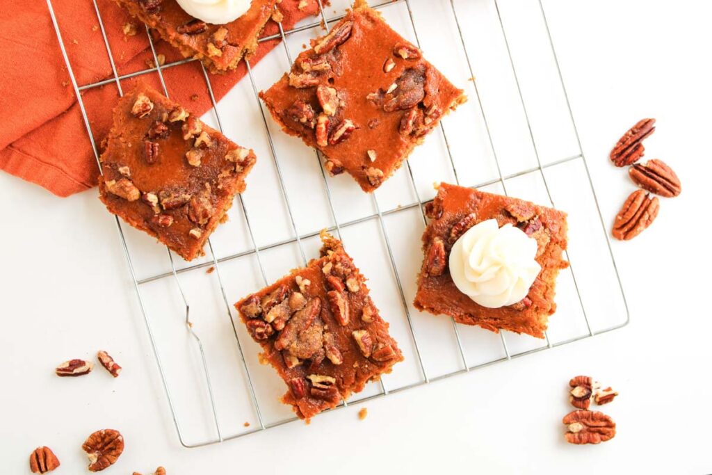 Pumpkin Pecan Pie Bars topped with cream and pecans are cooling on a wire rack, surrounded by scattered pecans and an orange napkin.