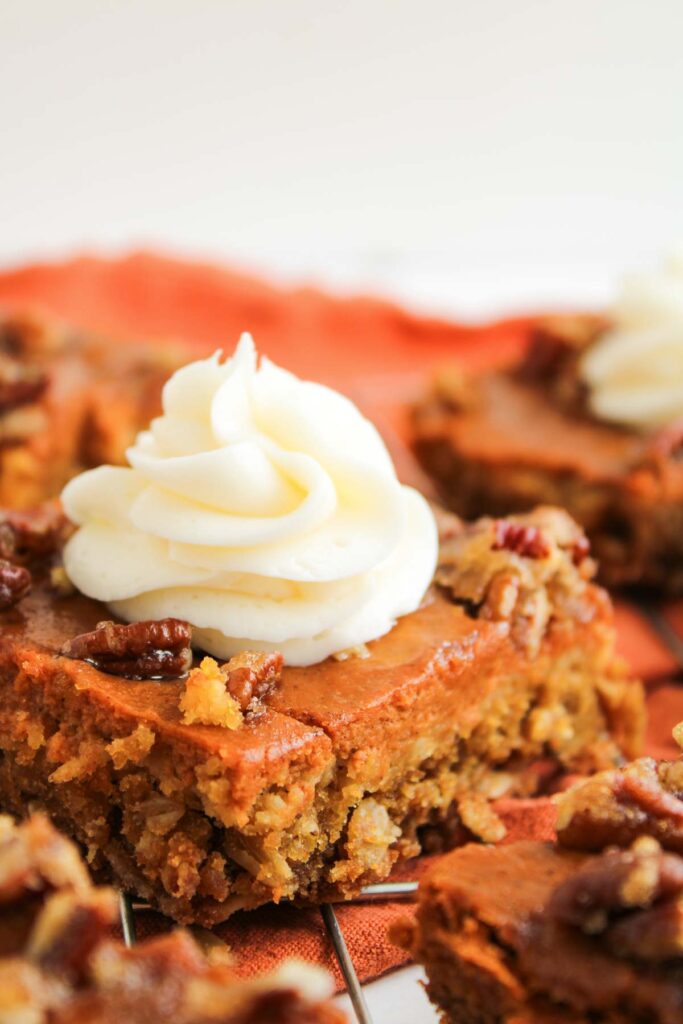 A close-up of Pumpkin Pecan Pie Bars, topped with a swirl of white frosting and chopped pecans, resting on a wire rack.