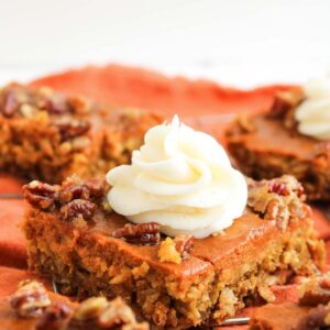 A close-up of Pumpkin Pecan Pie Bars topped with whipped cream and chopped pecans, placed on an orange cloth.