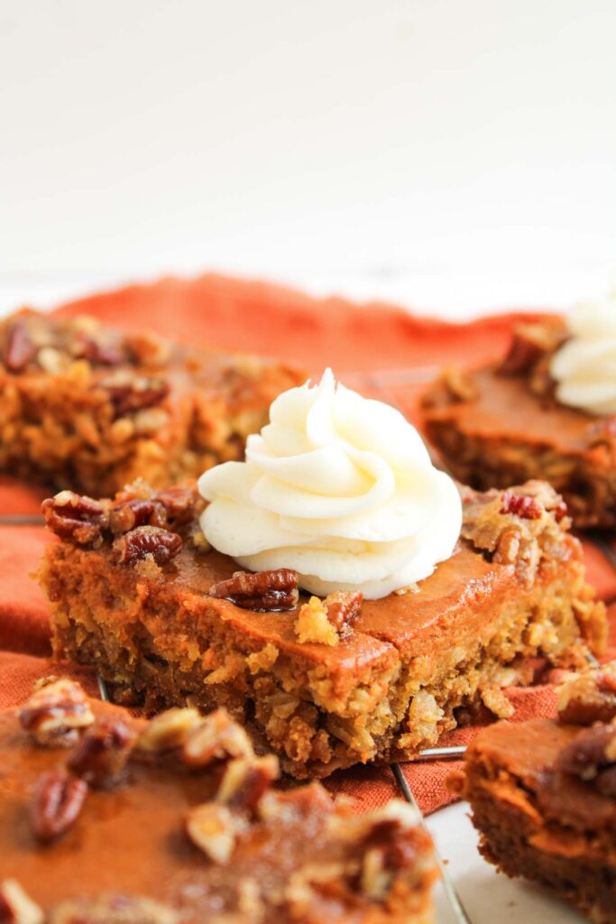 A close-up of Pumpkin Pecan Pie Bars topped with whipped cream and chopped pecans, placed on an orange cloth.