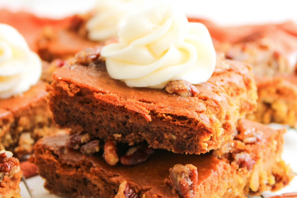 Two stacked Pumpkin Pecan Pie Bars topped with chopped nuts and swirled white frosting, with more bars visible in the background.