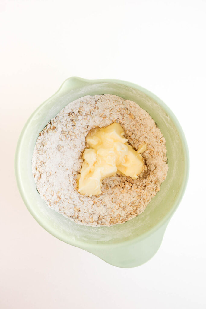 A green mixing bowl containing flour, oats, and a portion of butter placed on top—perfect for starting your Pumpkin Pecan Pie Bars—viewed from above.