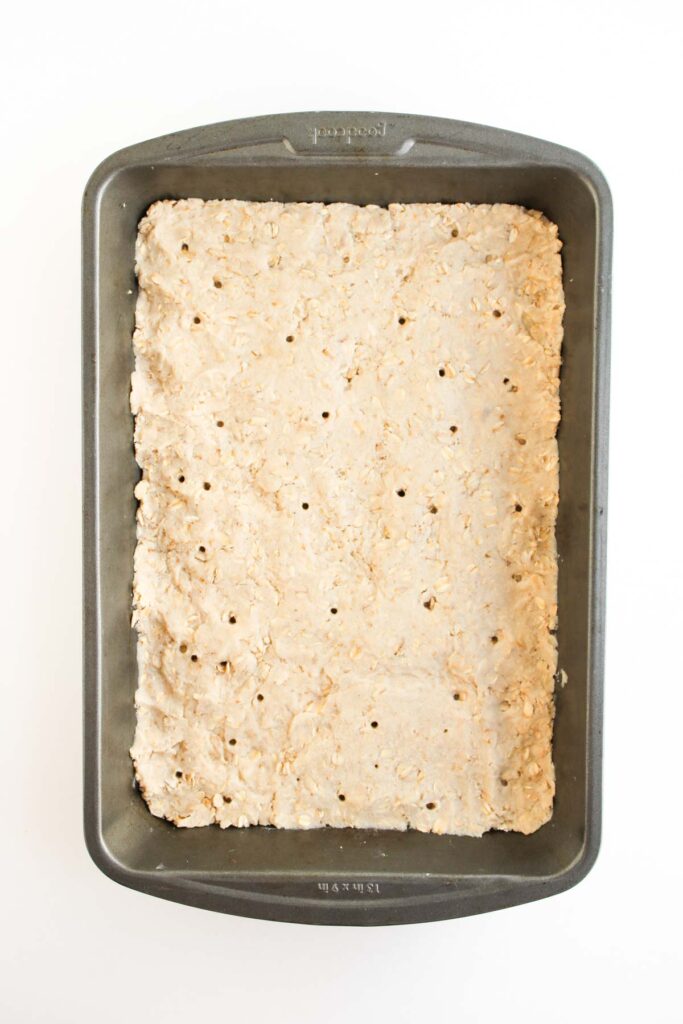 A rectangular metal baking pan filled with an unbaked oat crust dough, evenly spread and pricked with holes, ready to become the base for delicious Pumpkin Pecan Pie Bars.