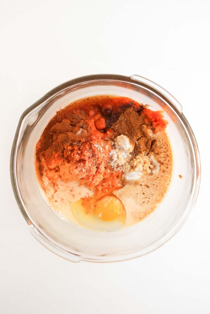 A glass mixing bowl contains ingredients for Pumpkin Pecan Pie Bars: an egg, pumpkin puree, milk, sugar, and various spices on a white surface.