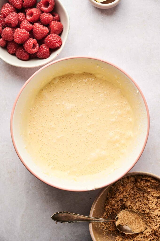 Large mixing bowl with pale batter for Air Fryer Raspberry Cheesecake, surrounded by a bowl of fresh raspberries and a bowl of brown sugar on a light surface.