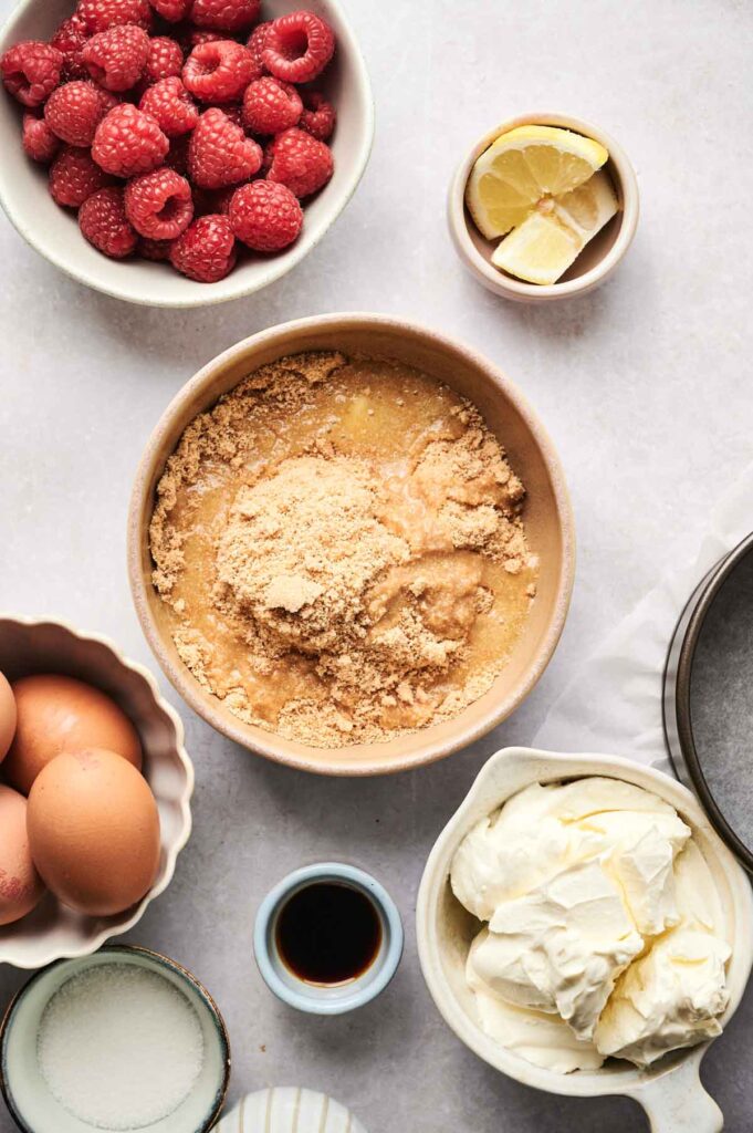 A bowl of crushed biscuits and melted butter sits among ingredients for an Air Fryer Raspberry Cheesecake, including fresh raspberries, lemon wedges, eggs, sugar, vanilla extract, and whipped cream cheese.