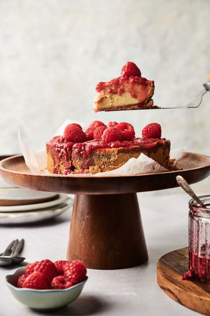 A slice of Air Fryer Raspberry Cheesecake is being lifted from a whole cheesecake on a wooden cake stand, with a bowl of raspberries and a jar of raspberry sauce nearby.