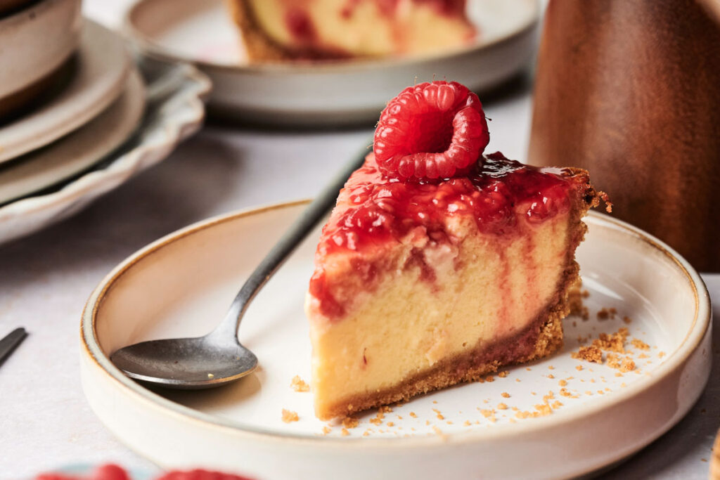 A slice of Air Fryer Raspberry Cheesecake on a plate with a spoon, topped with raspberry sauce and a fresh raspberry.