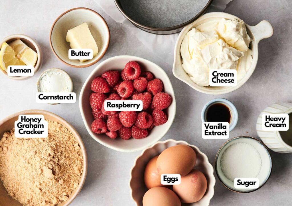 Various labeled bowls display ingredients for a delicious Air Fryer Raspberry Cheesecake: lemon, cornstarch, butter, graham cracker crumbs, raspberries, cream cheese, vanilla extract, heavy cream, eggs, and sugar.