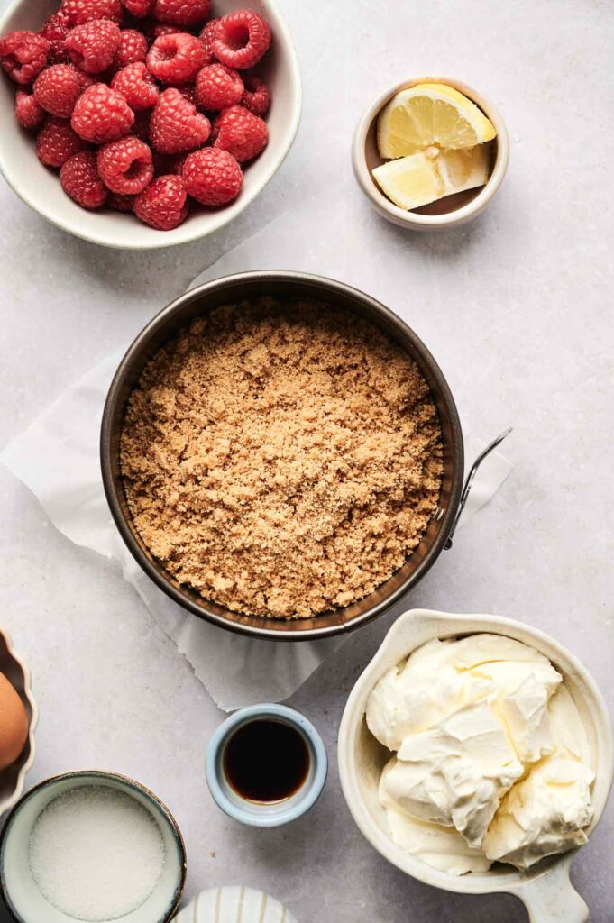 Overhead view of bowls containing ingredients for Air Fryer Raspberry Cheesecake&mdash;raspberries, lemon wedges, crushed graham crackers, sugar, vanilla extract, and whipped cream cheese on a light surface.