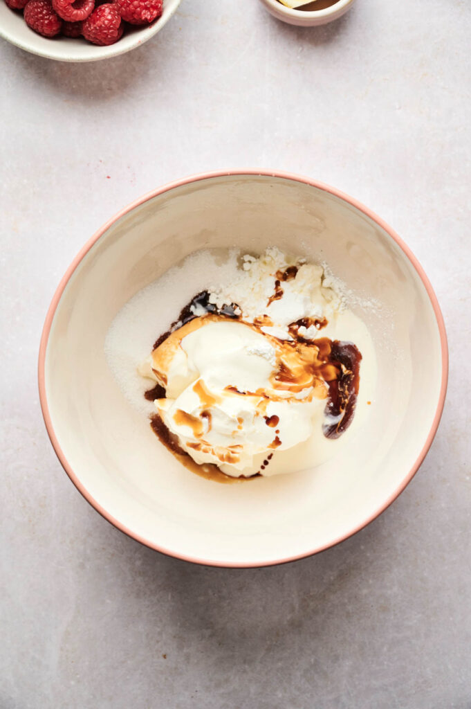 A mixing bowl containing sugar, sour cream, and vanilla extract&mdash;perfect for creating an Air Fryer Raspberry Cheesecake&mdash;with berries and a small bowl in the background.