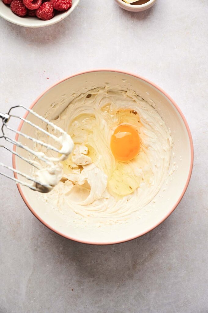 A mixing bowl contains creamed ingredients with a whole egg and egg yolk added, ready for the next step in making Air Fryer Raspberry Cheesecake; an electric mixer with batter on the beaters rests nearby.