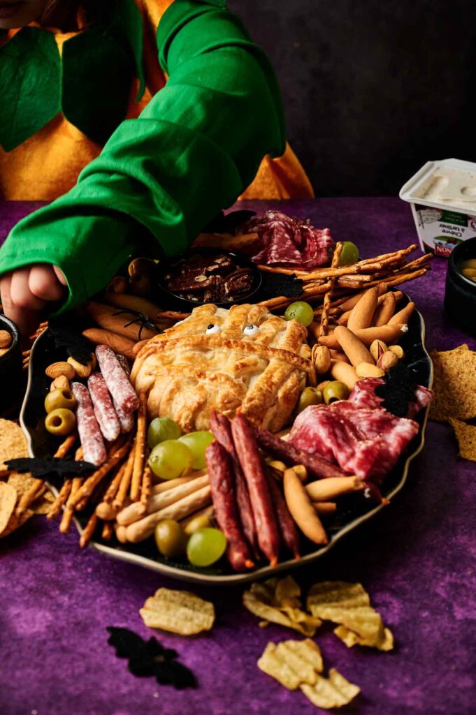 A festive charcuterie board with meats, breadsticks, grapes, olives, a pumpkin-shaped cheese ball, and a playful Camembert Cheese Mummy sits ready as a child in a green costume reaches for tasty treats.