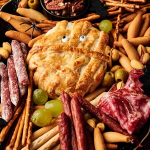 A charcuterie board with assorted meats, breadsticks, grapes, olives, nuts, and a playful Camembert Cheese Mummy pastry with candy eyes at the center.