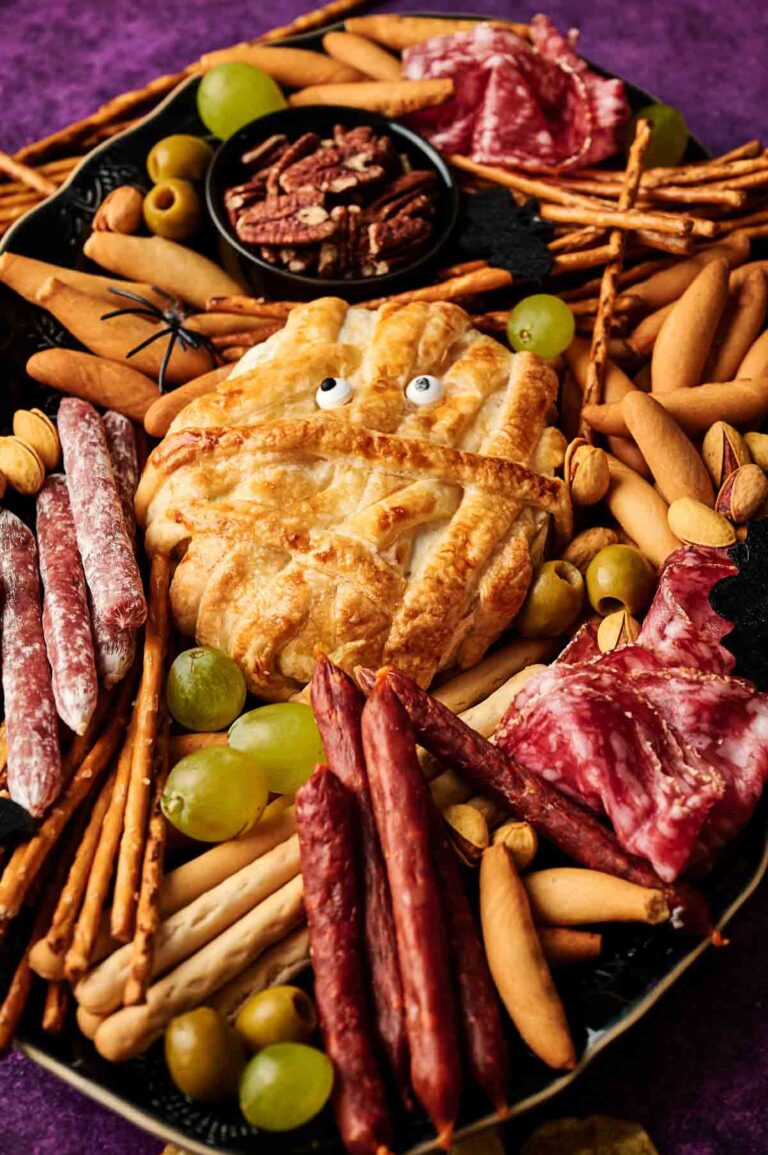 A charcuterie board with assorted meats, breadsticks, grapes, olives, nuts, and a playful Camembert Cheese Mummy pastry with candy eyes at the center.