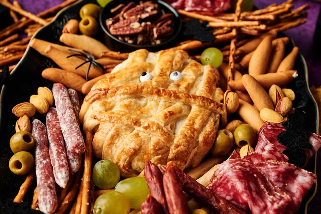 A Camembert Cheese Mummy appetizer with candy eyes is wrapped in flaky pastry and surrounded by cured meats, breadsticks, crackers, grapes, olives, and nuts on a black platter.