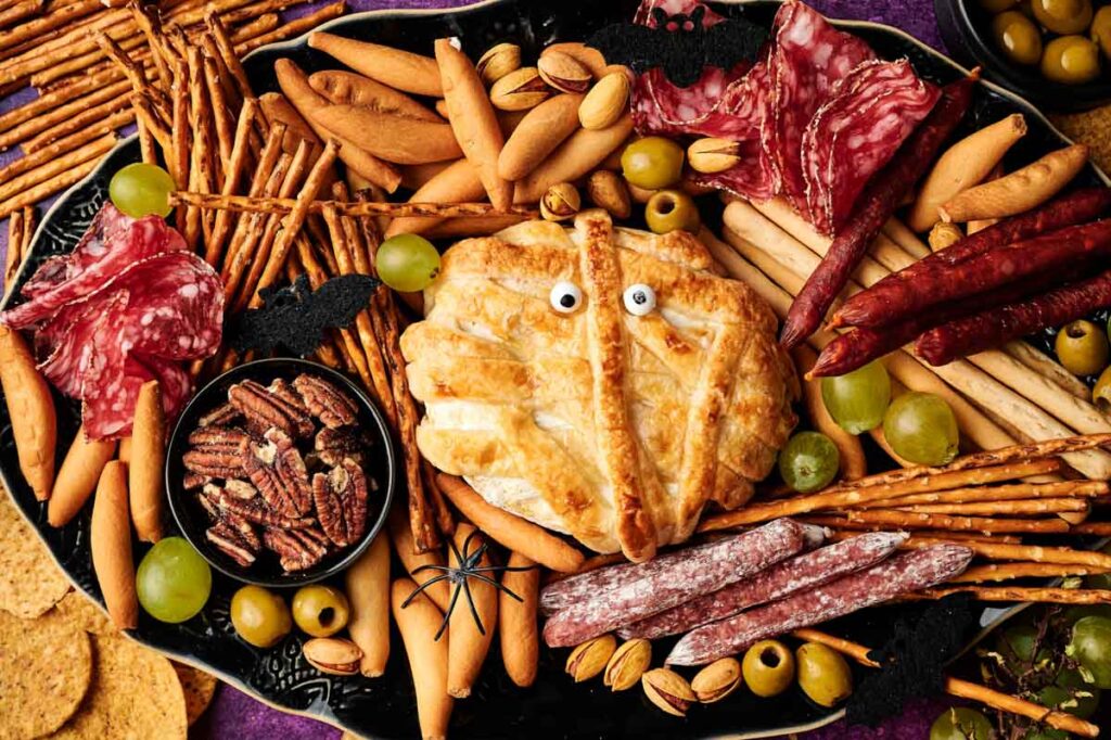 A Halloween-themed charcuterie board featuring assorted meats, breadsticks, pretzels, nuts, green grapes, and a spooky Camembert Cheese Mummy with candy eyes, all decorated with a playful plastic spider.