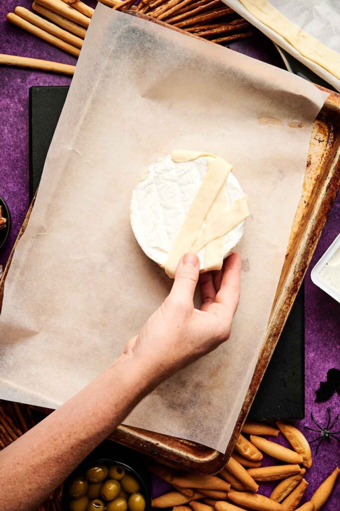 A hand places strips of dough over a round of brie, creating a Camembert Cheese Mummy on a parchment-lined baking sheet, with breadsticks and olives nearby.