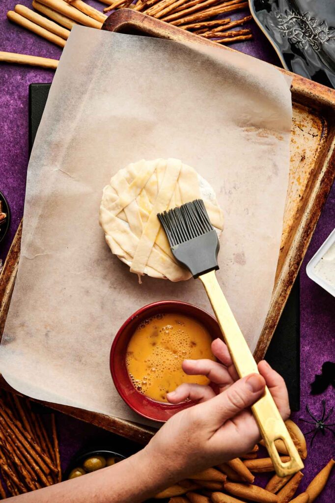 A hand uses a pastry brush to apply egg wash to a Camembert Cheese Mummy&mdash;round pastry wrapped with strips of dough&mdash;placed on a parchment-lined baking sheet.