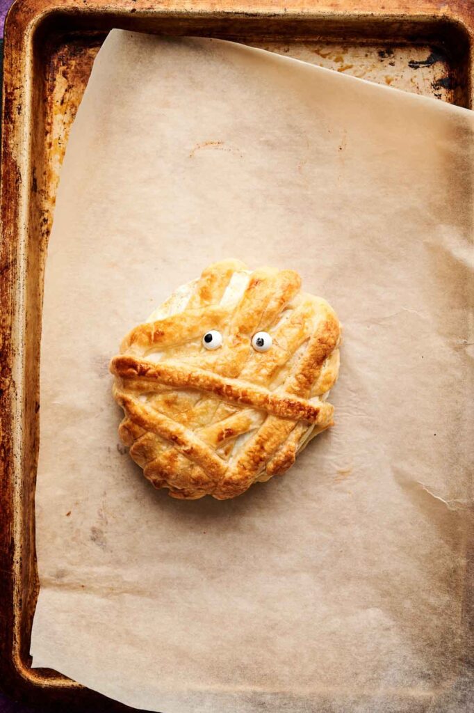 A round Camembert Cheese Mummy pastry decorated to look like a mummy, complete with two candy eyes, rests on parchment paper on a baking sheet.
