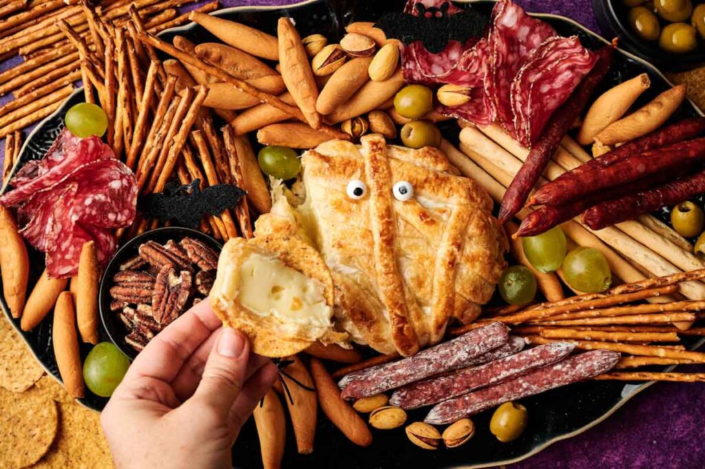 A hand lifts a piece of baked Camembert Cheese Mummy from a round pastry with candy eyes, surrounded by crackers, pretzels, meats, olives, nuts, and breadsticks on a platter.