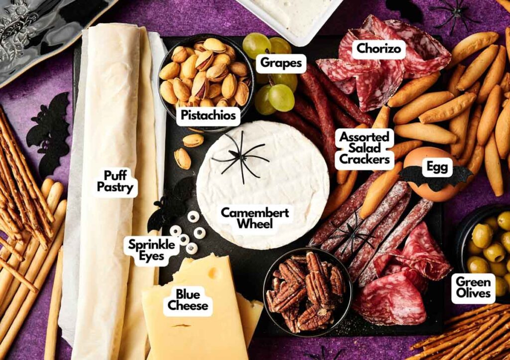 A Halloween-themed charcuterie board features a Camembert Cheese Mummy, puff pastry, sprinkle eyes, blue cheese, pistachios, grapes, chorizo, assorted crackers, egg, green olives, and pecans&mdash;all perfectly labeled for spooky snacking.