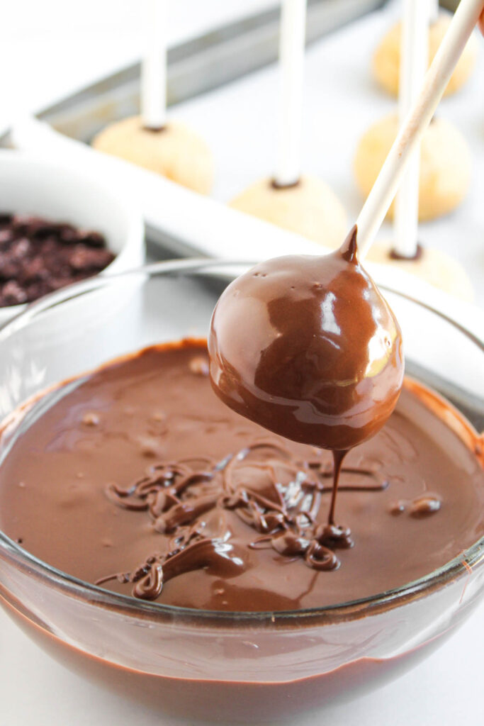A Worms and Dirt Cake Pop being dipped into a bowl of melted chocolate, with other cake pops and ingredients visible in the background.