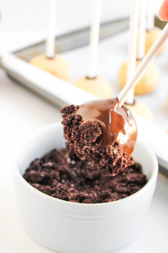 A Worms and Dirt Cake Pop is being dipped into a bowl of crushed chocolate cookies, with undipped cake pops on sticks visible in the background.